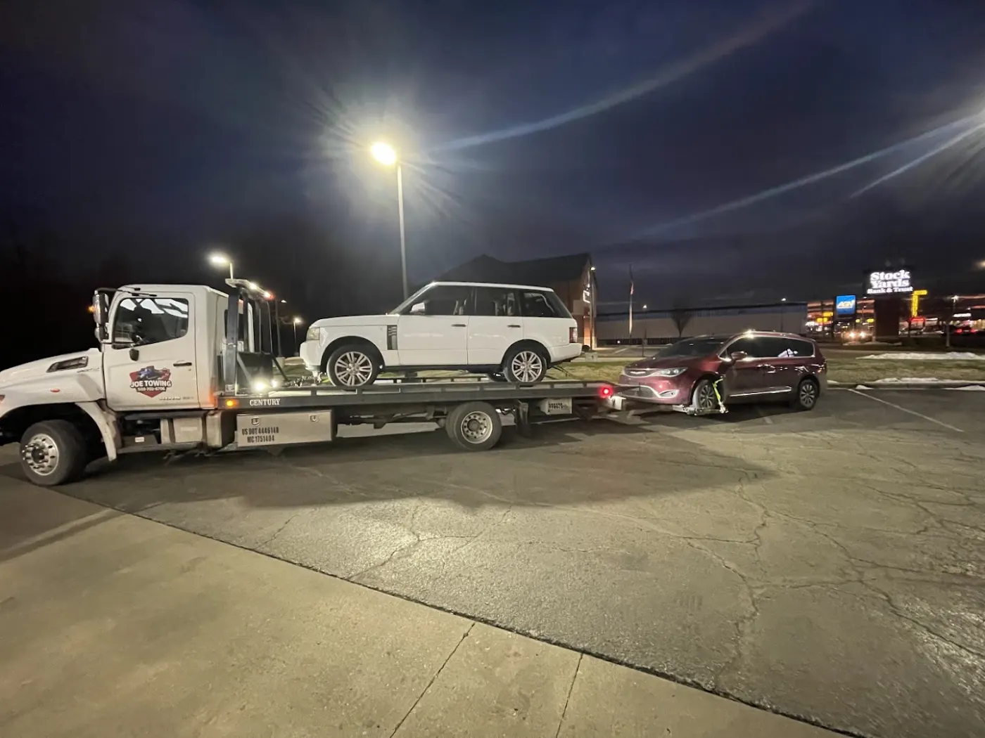 Joe Towing flatbed at night carrying a white SUV while towing a red minivan behind it.
