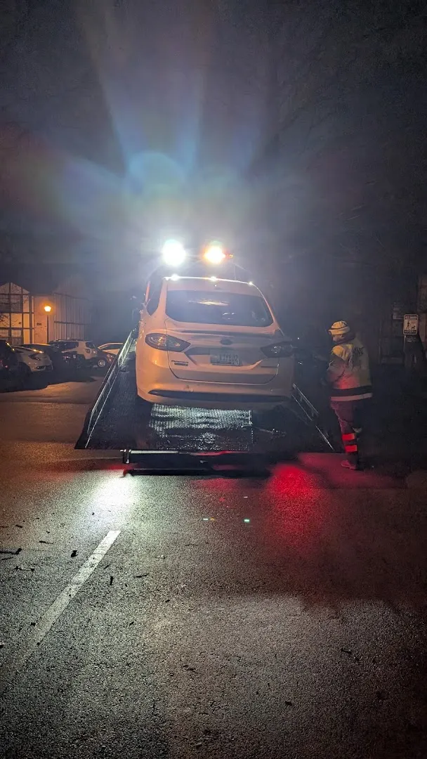 Tow operator in a reflective vest beside a white sedan loaded on the flatbed at night.